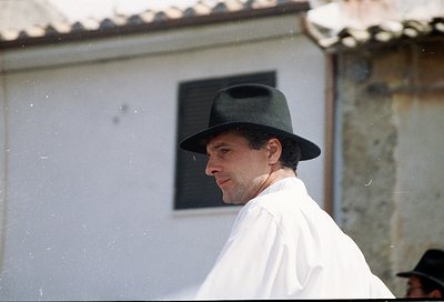 Portrait of a man in a wide-brimmed hat and light-colored robe, standing near a rustic stone building with a tiled roof. The ...