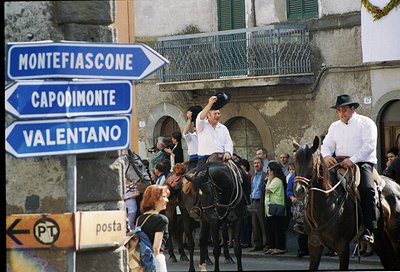 Two men in formal attire ride horses through a historic Italian town square, waving. Blue directional signs point to Montefia...