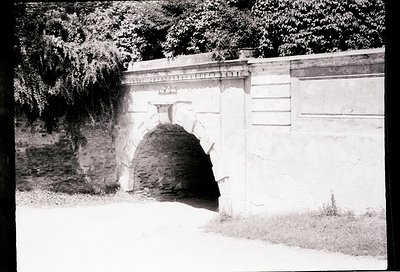Historic stone archway with weathered brick tunnel entrance, flanked by high concrete walls. Likely part of a fortification o...