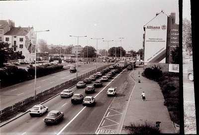 Mid-20th century urban road scene with heavy traffic, featuring vintage cars and a prominent **Allianz** billboard advertisin...