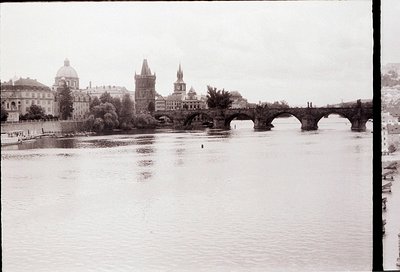 Vintage black-and-white view of Prague’s Charles Bridge spanning the Vltava River, flanked by historic towers and domes. Goth...