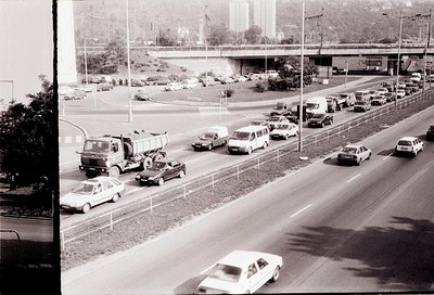 Mid-20th century urban highway jam with vintage vehicles, including a tow truck assisting a disabled car. Concrete barriers a...