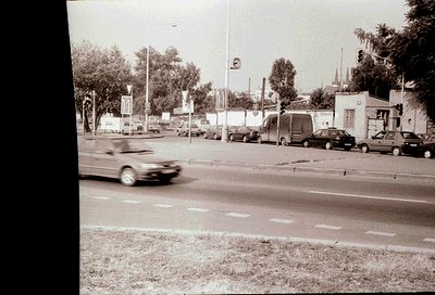 Urban street scene with motion blur of a passing car, lined with parked vehicles and greenery. Mid-20th century architecture ...