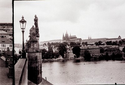 Black-and-white view of Prague’s **Charles Bridge** with ornate stone statues flanking the railing. Gothic spires of **Prague...