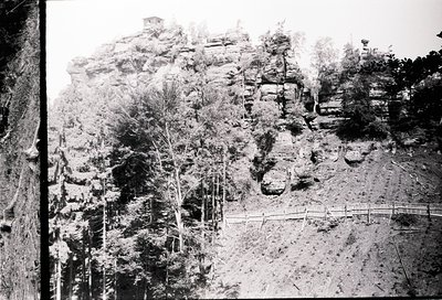 Early 20th-century black-and-white landscape of rugged rock formations and forested slopes. A wooden fence traces a path alon...