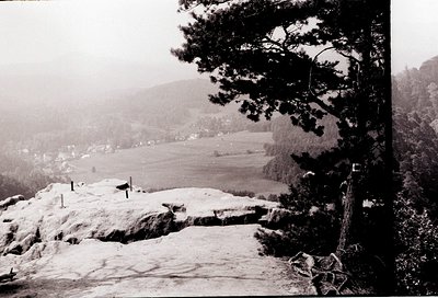 Black-and-white landscape featuring snow-covered rocky outcrop with a narrow path winding through. Dense forest frames right ...