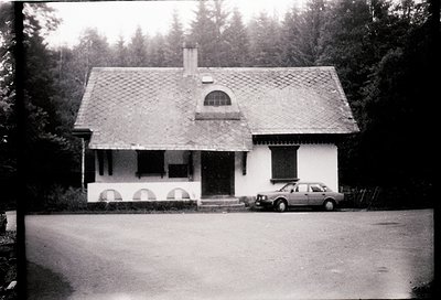 Classic mid-20th century alpine cottage with gabled roof and arched windows, set in forested surroundings. Vintage sedan park...