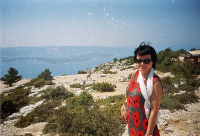 Vintage seaside portrait of woman in 1970s-style patterned dress, posing on rocky coastal terrain. Clear blue horizon with di...