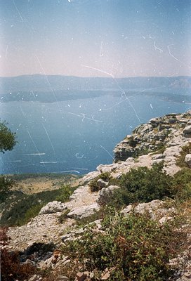 Vintage photograph showing a rugged coastal cliffside with sparse vegetation and a deep blue sea extending to distant landfor...