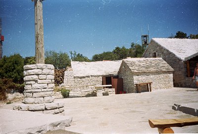 Rustic stone courtyard with traditional rural architecture—stone-built huts with pitched roofs, a wooden cross, and a stone p...