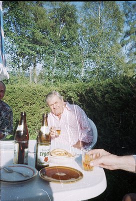 Mid-century outdoor dining scene featuring an older man in a light-colored shirt, seated at a round table with a bottle of *R...