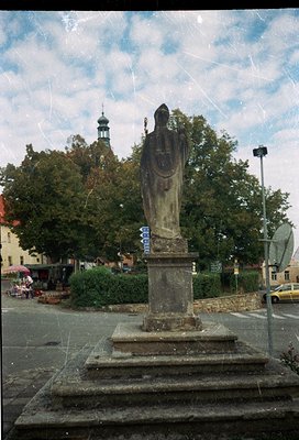 Stone statue of a robed figure holding a cross, mounted on a pedestal in an urban square. Surrounding elements include direct...