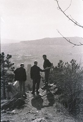 Three men in formal attire pose atop rugged terrain, overlooking a valley with distant hills. Mid-20th century black-and-whit...