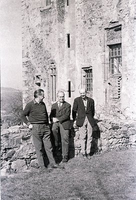 Three men pose outside a weathered stone building with arched windows, likely mid-20th century. Formal attire suggests a prof...