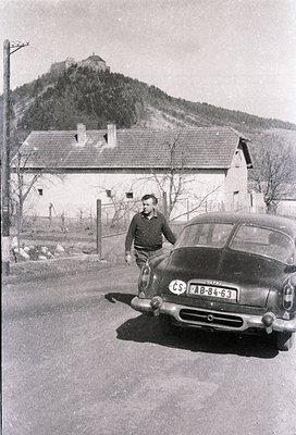 Mid-20th century black-and-white photo featuring a man in a dark jacket and cap standing beside a vintage **Škoda AB-84-63** ...