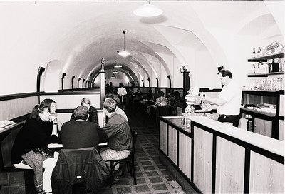 Vintage underground café with arched brick ceilings and tiled floors, featuring 1970s-style decor. Patrons seated at high tab...
