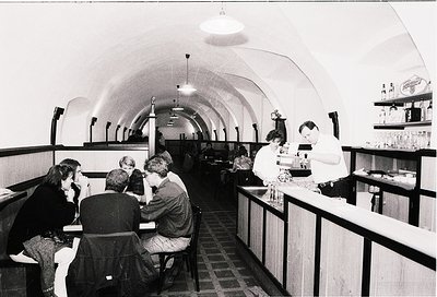 Vintage underground café with curved brick ceiling and exposed lighting fixtures. Patrons seated at wooden tables converse, w...
