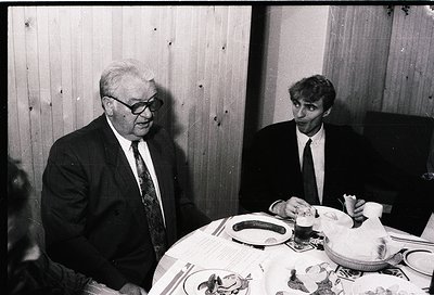 Two men in formal attire share a meal at a wooden-paneled table, likely mid-20th century. The older man wears glasses and a s...