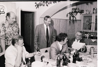 Mid-century indoor gathering in a modest kitchen/dining area, featuring five adults seated/standing around a table. Men wear ...