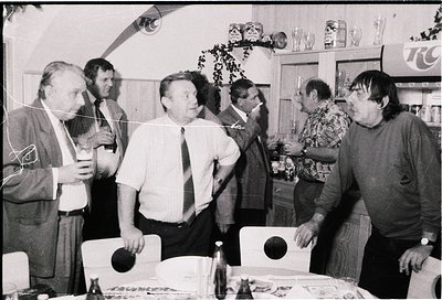 Group of six men in a 1970s-era indoor setting, likely a social gathering. Centered around a table with food, drinks (bottles...