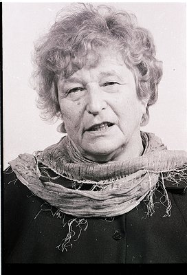 Portrait of an elderly woman in mid-speech, wearing a textured scarf and dark jacket. Black-and-white, high-contrast style su...