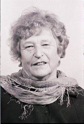 Portrait of an elderly woman in mid-20th century attire, likely 1950s–1960s. She wears a dark jacket with a frayed, textured ...