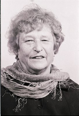 Portrait of an elderly woman with short, wavy gray hair, smiling directly at the camera. She wears a dark jacket with a textu...