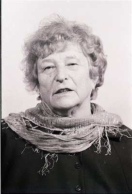 Black-and-white portrait of an elderly woman with short, curly white hair, wearing a textured scarf and dark coat. Expression...