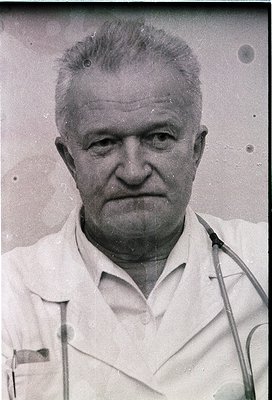 Mid-century black-and-white portrait of an older man in a white lab coat, holding a stethoscope. The grainy, aged film sugges...