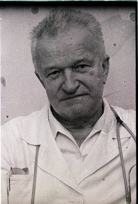 Mid-century black-and-white portrait of an elderly man in a white lab coat, likely a physician or scientist. His expression i...