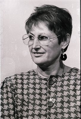 Portrait of a woman in a patterned blouse with geometric checkered design, wearing round-framed glasses and large hoop earrin...
