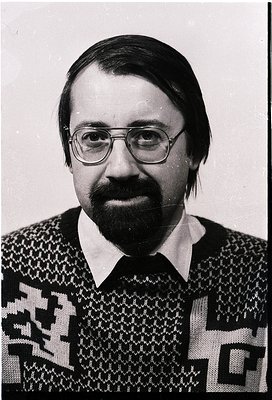 Portrait of a man in a patterned sweater with geometric motifs, likely 1970s-1980s. Glasses and short beard frame a neutral e...