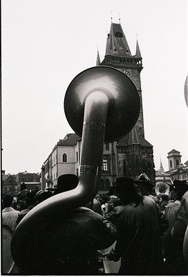 Black-and-white street scene featuring a brass band’s tuba in focus, with a Gothic-style church spire (likely Prague’s Old To...