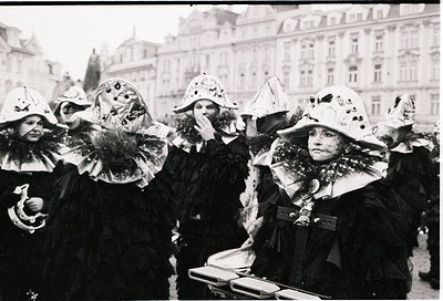 Black-and-white street scene featuring women in elaborate, ornate carnival masks and fur-trimmed coats, likely from a 19th–ea...