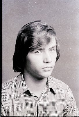 Portrait of a young man in a plaid button-up shirt, likely a 1970s school or ID photo. Neutral expression, short side-swept h...