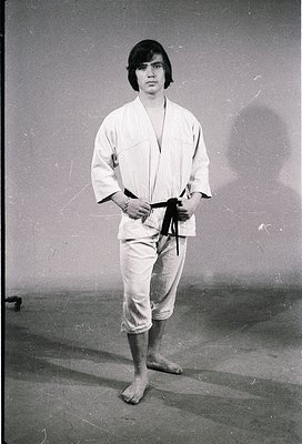 Portrait of a person in traditional judo attire (gi), standing indoors with neutral background. Belt appears to be black or d...