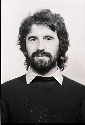 Portrait of a man with long sideburns, mustache, and beard, wearing a dark sweater and striped collared shirt. Classic 1970s-...