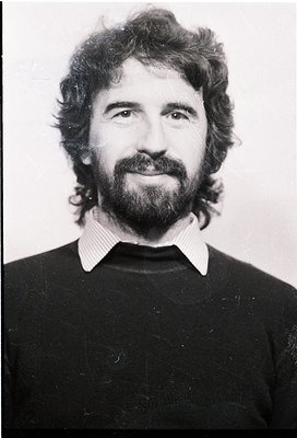 Portrait of a man with a full beard and mustache, wearing a striped collared shirt under a dark sweater. Mid-length, wavy hai...