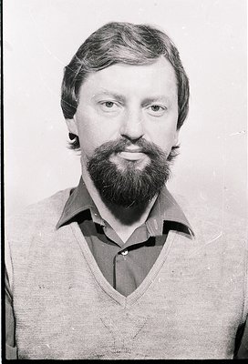 Portrait of a man with a full beard and mustache, wearing a V-neck sweater. Style suggests 1970s–1980s Western Europe/USA. Ne...