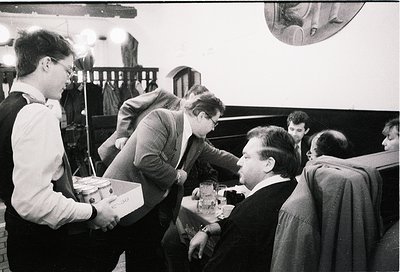 Vintage black-and-white photo of a group of men in formal attire (suits, ties) playing chess in a dimly lit room. One man in ...