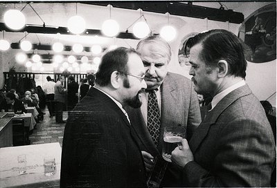 Three men in formal attire—dark suits, ties, and dress shirts—engage in conversation at a mid-20th-century indoor event. Susp...