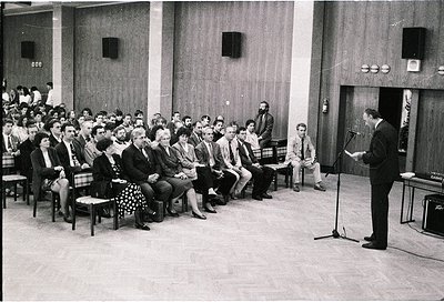 Mid-century lecture hall with tiered seating, wooden paneling, and industrial lighting. A speaker addresses an attentive audi...