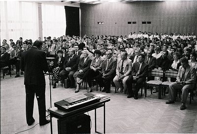 A formal lecture or conference in a mid-century auditorium, featuring a speaker addressing a packed audience seated in tiered...