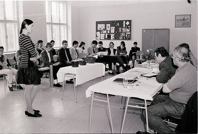 Black-and-white classroom scene featuring a teacher addressing students seated at long tables, 1960s–1970s. Functional, insti...