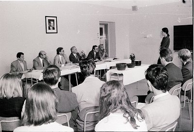 Black-and-white conference scene: seated panelists at a rectangular table in a plain room, with one speaker addressing an aud...
