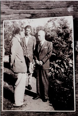 Three men in formal 1950s–60s attire pose outdoors in a lush garden setting. The older man in a dark suit and tie gestures to...