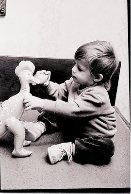 Mid-20th century black-and-white photo of a young child playing with a large ceramic duck toy, seated on a carpeted floor. Th...
