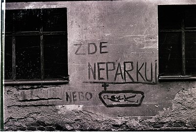 Black-and-white graffiti on weathered concrete wall reads *"ZDE NEPARKUJ!"* ("Don’t park here!") with a crossed-out parking s...