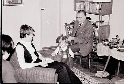 Black-and-white interior shot of a mid-20th-century home, likely 1960s–1970s. A man in formal attire (vest, tie) interacts wi...