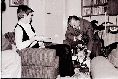 Mid-20th century living room scene: a woman in a turtleneck sweater holds a tray, an elderly man in suit and tie interacts wi...